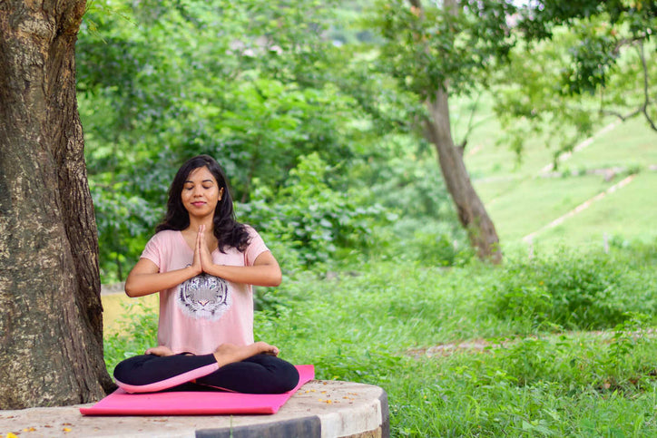 Woman meditating in a seated yoga pose outdoors near a tree.